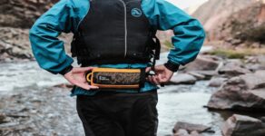 Man in blue water jacket and life vest wearing a orange River Station fanny pack, standing on a river in the mountains.