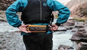 Man in blue water jacket and life vest wearing a orange River Station fanny pack, standing on a river in the mountains.