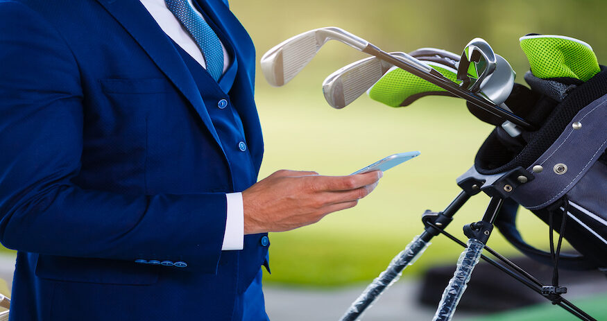 Man wearing a blue suit doing business on the golf course while on his phone, next to his golf bag.