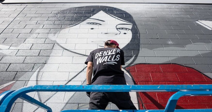 man painting on a city wall, wearing a black tee-shirt that says "DENVER WALLS"