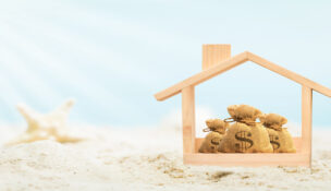 Wooden home model and money bag put on the beautiful sand and starfish on blue sky background in summer vacation, Saving or loan for buy a new house or real estate owner concept.