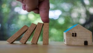 The hand stops wooden dominoes falling to house model, real estate protection concept background. Hands stop the domino effect before destroying a home.