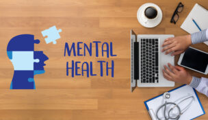 Mental Health in the workplace concept: Man typing on his computer with a messy desk and the words "MENTAL HEALTH" displayed next to a puzzle depicting a human head.