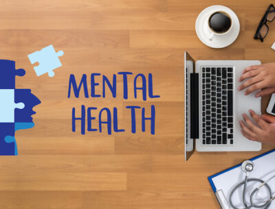 Mental Health in the workplace concept: Man typing on his computer with a messy desk and the words "MENTAL HEALTH" displayed next to a puzzle depicting a human head.